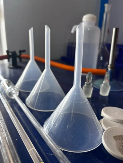 Three plastic filter funnels beside glass stoppers, glass tubes and white porcelain dishes on a blue bench