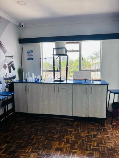 Long lab bench marked Physics under a window, with a sink, measuring cylinders, gas taps and a small whiteboard