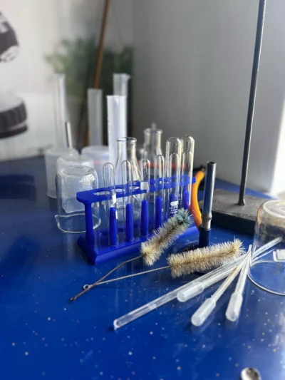 Blue test tube rack with test tubes, two test tube brushes, plastic droppers and beakers on a blue bench